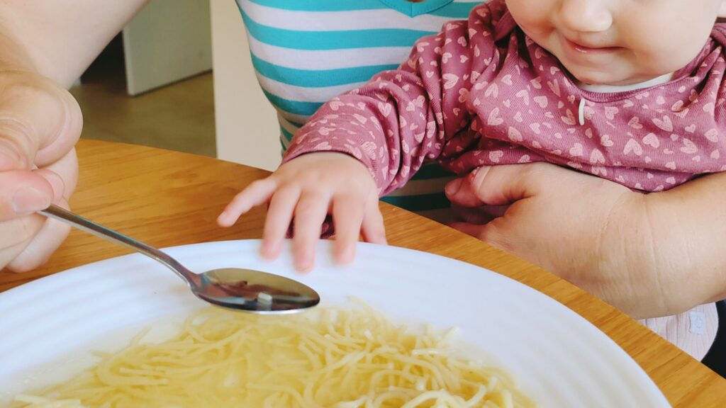 Matka karmiąca dziecko rosołem, podczas gdy dziecko siedzi na jej kolanach. Scena pełna ciepła i rodzinnej atmosfery, podkreślająca tradycję wspólnego posiłku i zdrowego odżywiania.